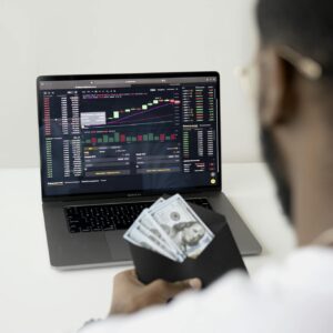 A person examines stock market data on a laptop while holding dollar bills, showcasing finance and trading concepts.