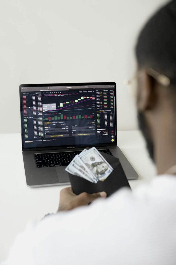 A person examines stock market data on a laptop while holding dollar bills, showcasing finance and trading concepts.